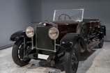 A vintage car with large round headlights, a dark sleek body, and a convertible top. The vehicle is parked indoors on a marble floor and has classic design elements with a visible spare tire attached to the side.