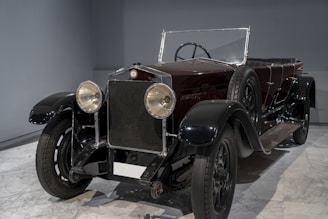 A vintage car with large round headlights, a dark sleek body, and a convertible top. The vehicle is parked indoors on a marble floor and has classic design elements with a visible spare tire attached to the side.