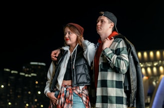 A young couple is standing together at night with city lights in the background. Both are wearing casual clothing with jackets and shirts. The woman has her arm around the man's shoulders, and he has his hand in his pocket.
