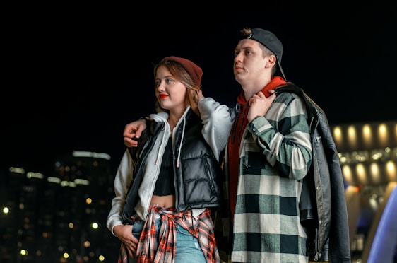 A young couple is standing together at night with city lights in the background. Both are wearing casual clothing with jackets and shirts. The woman has her arm around the man's shoulders, and he has his hand in his pocket.