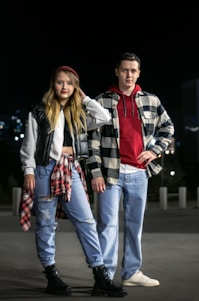 A young man and woman stand confidently in an urban environment at night. They are both dressed in casual attire with a trendy, layered look. The woman wears ripped jeans, a black vest, and a plaid shirt tied around her waist. The man wears jeans, a red hoodie, and a checkered shirt over it. They have a youthful, relaxed demeanor.