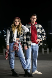 A young man and woman stand confidently in an urban environment at night. They are both dressed in casual attire with a trendy, layered look. The woman wears ripped jeans, a black vest, and a plaid shirt tied around her waist. The man wears jeans, a red hoodie, and a checkered shirt over it. They have a youthful, relaxed demeanor.