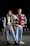A young man and woman stand confidently in an urban environment at night. They are both dressed in casual attire with a trendy, layered look. The woman wears ripped jeans, a black vest, and a plaid shirt tied around her waist. The man wears jeans, a red hoodie, and a checkered shirt over it. They have a youthful, relaxed demeanor.