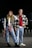 A young man and woman stand confidently in an urban environment at night. They are both dressed in casual attire with a trendy, layered look. The woman wears ripped jeans, a black vest, and a plaid shirt tied around her waist. The man wears jeans, a red hoodie, and a checkered shirt over it. They have a youthful, relaxed demeanor.