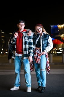Two individuals stand casually outdoors at night with a modern building illuminated in the background. They are dressed in casual attire, including jeans and plaid shirts, with one person wearing a hat and the other with a tied plaid shirt around the waist.