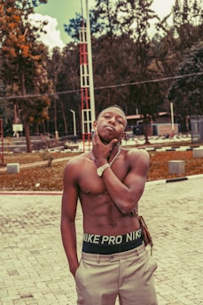 A shirtless young man stands confidently in a park-like area with a chain bracelet on his wrist and tailored pants. His expression is relaxed yet assertive, with his chin resting on his left hand. There are trees and a red and white tower in the background.