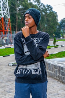 A young person wearing a beanie and a black sweatshirt stands outdoors, with a thoughtful expression, resting their hand on their chin. The background features green trees, a paved area, and a large metal structure.