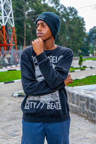 A young person wearing a beanie and a black sweatshirt stands outdoors, with a thoughtful expression, resting their hand on their chin. The background features green trees, a paved area, and a large metal structure.