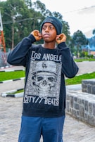 A person is adjusting their earphones outdoors. They are wearing a black beanie and a dark sweatshirt with a graphic design featuring a skull and the words 'Los Angeles City Beats'. The background includes greenery and concrete structures.