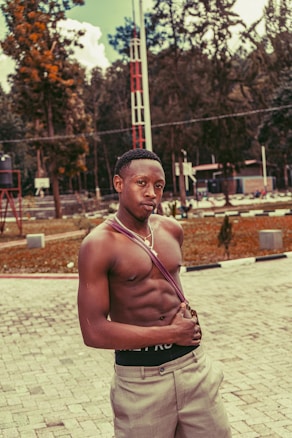 A shirtless man stands confidently in an outdoor setting with a path made of stone tiles and tall trees in the background. He is wearing light-colored pants and a cross-body strap, with his gaze directed towards the camera.