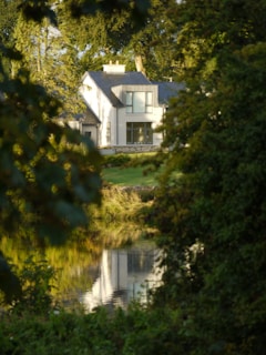 A modern two-story house is partially visible through dense foliage, with large windows reflecting sunlight. The house is situated near a calm body of water that mirrors its image. Tall trees surround the area, suggesting a secluded, serene setting.