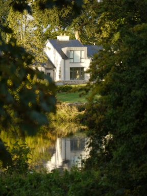 A modern two-story house is partially visible through dense foliage, with large windows reflecting sunlight. The house is situated near a calm body of water that mirrors its image. Tall trees surround the area, suggesting a secluded, serene setting.