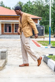 A man is dressed in a tan blazer, beige trousers, and brown shoes, walking on a paved path beside a building with a tiled roof and red brick exterior. Trees can be seen in the background.