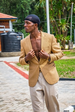 A person wearing a beige blazer and light-colored pants stands in an outdoor setting. The individual is adjusting the blazer with a confident pose. In the background, there is greenery and a brick structure with a red roof, along with a large black water tank.