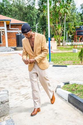 A person wearing a tan blazer, light-colored trousers, and brown shoes walks along a paved path in an outdoor setting. The background features a brick building with a red roof and lush greenery, including tall trees and shrubs.