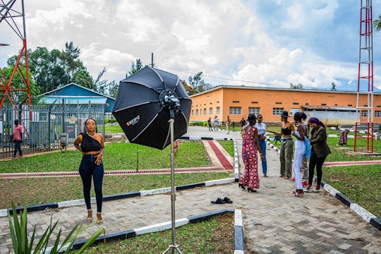 A woman stands confidently in a photoshoot setup outdoors, with a large professional umbrella lighting equipment beside her. In the background, a group of people are gathered, engaging in conversation. This urban setting features a grassy area with pathways and utility buildings, as well as a tall metal structure.