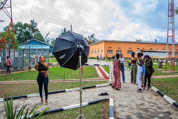 A woman stands confidently in a photoshoot setup outdoors, with a large professional umbrella lighting equipment beside her. In the background, a group of people are gathered, engaging in conversation. This urban setting features a grassy area with pathways and utility buildings, as well as a tall metal structure.