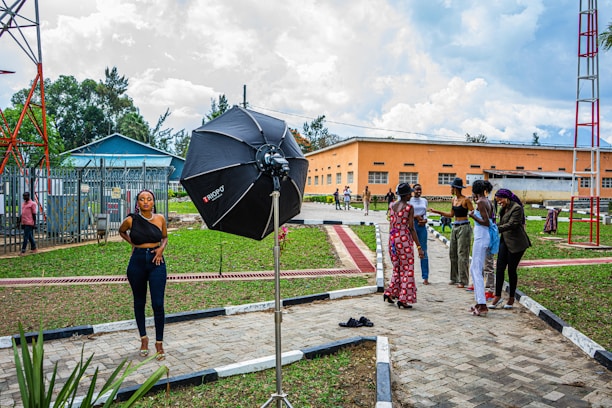A woman stands confidently in a photoshoot setup outdoors, with a large professional umbrella lighting equipment beside her. In the background, a group of people are gathered, engaging in conversation. This urban setting features a grassy area with pathways and utility buildings, as well as a tall metal structure.