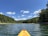 A kayak glides across a calm lake surrounded by lush green forests under a bright blue sky with scattered white clouds. The perspective from the bow of the kayak gives a sense of exploration and adventure.