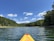A kayak glides across a calm lake surrounded by lush green forests under a bright blue sky with scattered white clouds. The perspective from the bow of the kayak gives a sense of exploration and adventure.