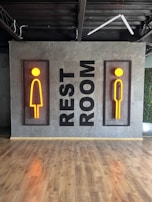 A modern restroom sign featuring stylized male and female figures in vibrant orange, mounted on a textured gray wall. The words 'REST ROOM' are displayed vertically between the two signs. The floor is made of polished wood, and there's visible industrial ceiling infrastructure.