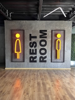A modern restroom sign featuring stylized male and female figures in vibrant orange, mounted on a textured gray wall. The words 'REST ROOM' are displayed vertically between the two signs. The floor is made of polished wood, and there's visible industrial ceiling infrastructure.