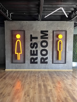 A modern restroom sign featuring stylized male and female figures in vibrant orange, mounted on a textured gray wall. The words 'REST ROOM' are displayed vertically between the two signs. The floor is made of polished wood, and there's visible industrial ceiling infrastructure.