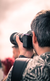A person with short hair holds a camera, capturing a scene. The focus is on the camera and the person's hand wrapped around it. Background is softly blurred and slightly overexposed, creating a dreamy atmosphere.