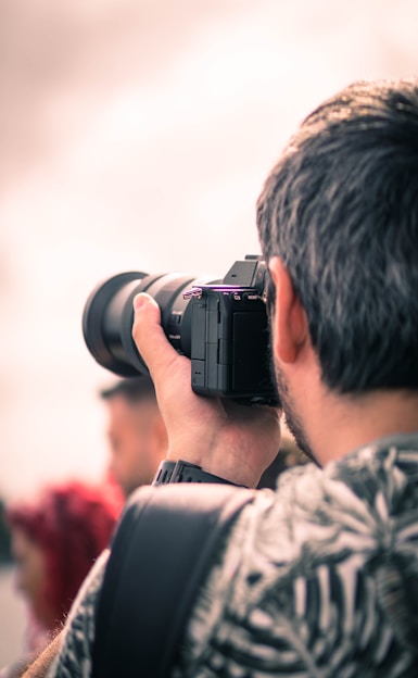 A person with short hair holds a camera, capturing a scene. The focus is on the camera and the person's hand wrapped around it. Background is softly blurred and slightly overexposed, creating a dreamy atmosphere.