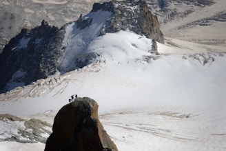 A group of climbers is reaching the summit of a rocky peak amidst a vast, snow-covered mountainous landscape. The background shows rugged terrain with patches of snow and exposed rock formations.
