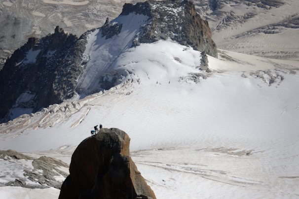 A group of climbers is reaching the summit of a rocky peak amidst a vast, snow-covered mountainous landscape. The background shows rugged terrain with patches of snow and exposed rock formations.