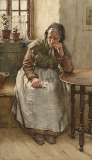 An elderly woman sits pensively in a modest, dimly lit room. She wears a shawl and apron, her expression thoughtful as she rests her chin on one hand. A wooden table is beside her, and a window with potted plants allows light to filter through, casting soft shadows.