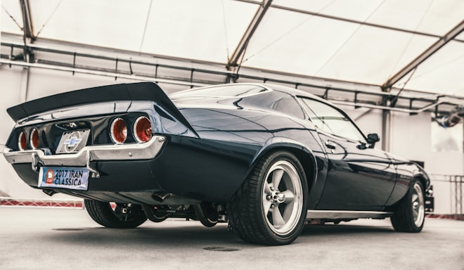 A classic muscle car is parked in a well-lit indoor setting, showcasing its sleek lines and powerful build. The vehicle features a glossy dark blue paint job, prominent rear spoiler, and polished chrome elements. The wheels are large and sporty, complementing the car's vintage design with a modern touch. A license plate indicates participation in a classic car show in 2017.
