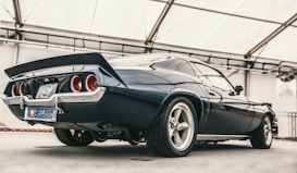A classic muscle car is parked in a well-lit indoor setting, showcasing its sleek lines and powerful build. The vehicle features a glossy dark blue paint job, prominent rear spoiler, and polished chrome elements. The wheels are large and sporty, complementing the car's vintage design with a modern touch. A license plate indicates participation in a classic car show in 2017.