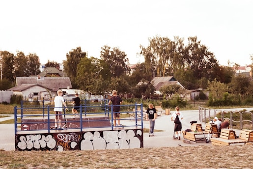 A skate park scene with several young people engaging in various activities. A few of them are on skateboards inside a small fenced area with graffiti on the side. Others are sitting on benches, possibly resting or watching. The background includes trees and houses under a clear sky.