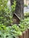 A small wooden insect house is nestled among lush green foliage and leaves, positioned against the trunk of a large tree. Surrounding the setup is a wooden fence, all contributing to a natural, serene garden setting.