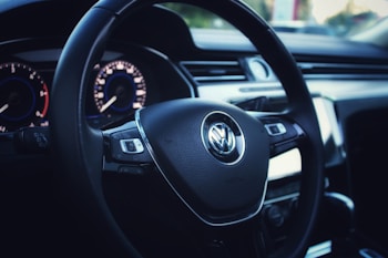 A close-up view of a Volkswagen car steering wheel, showcasing the brand logo prominently in the center. The dashboard behind the steering wheel displays illuminated dials with blue and red accents. The interior has a modern and sleek design with a polished look.