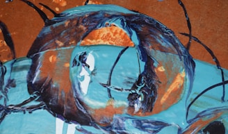 An abstract painting featuring a swirling combination of blue, orange, and black colors. Thick, textured brushstrokes create dynamic circular patterns on a brown background, giving a sense of motion and depth.