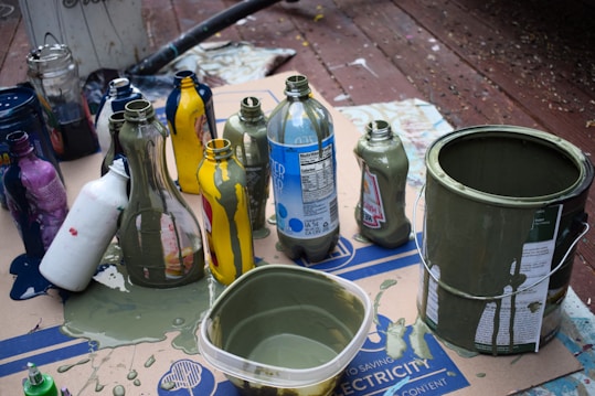 Various recycled bottles and containers covered in paint are arranged on a wooden surface. The bottles are different shapes and colors, with visible paint drips and splashes of green and blue paint on them. A paint can and a plastic container, both filled with green paint, are also present on a piece of cardboard beneath them.