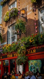 A lively street scene in front of a popular bar during the daytime. The upper portion of the bar is adorned with vibrant flowerboxes, hanging plants, and two illuminated lanterns. The establishment has a red facade with 'The Temple Bar' sign prominently displayed. There are various signs and posters on the bar’s walls, including a Guinness sign, and a painting of a man with the text 'The Temple Bar' in the window. A crowd of people is gathered outside, some engaged in conversation, others walking by.