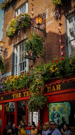 A lively street scene in front of a popular bar during the daytime. The upper portion of the bar is adorned with vibrant flowerboxes, hanging plants, and two illuminated lanterns. The establishment has a red facade with 'The Temple Bar' sign prominently displayed. There are various signs and posters on the bar’s walls, including a Guinness sign, and a painting of a man with the text 'The Temple Bar' in the window. A crowd of people is gathered outside, some engaged in conversation, others walking by.