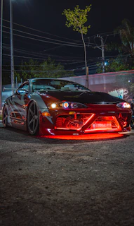 A sleek JDM car with vibrant decals parked at a bustling car meet under evening lights