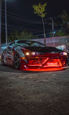 A sports car with a sleek, aerodynamic design is parked on a street at night. It has a custom body kit, large alloy wheels, and vibrant red underglow lights. The car is illuminated under the streetlights, and the background shows trees, power lines, and other urban elements.