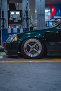A dark green car is parked at a gas station. The focus is on the front left wheel, which has a sleek, metallic rim design. Two fuel pumps with hoses are in the background, and various signs and indicators are present on the pumps.