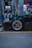 A dark green car is parked at a gas station. The focus is on the front left wheel, which has a sleek, metallic rim design. Two fuel pumps with hoses are in the background, and various signs and indicators are present on the pumps.