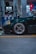 A dark green car is parked at a gas station. The focus is on the front left wheel, which has a sleek, metallic rim design. Two fuel pumps with hoses are in the background, and various signs and indicators are present on the pumps.