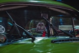 A close-up view of the windshield of a vintage green car, featuring two windshield wipers and a hanging air freshener in the shape of a car. The interior is partially visible, showing a red safety belt. Various decals are present on the glass, including a club sticker.