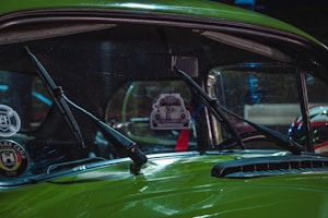 A close-up view of the windshield of a vintage green car, featuring two windshield wipers and a hanging air freshener in the shape of a car. The interior is partially visible, showing a red safety belt. Various decals are present on the glass, including a club sticker.