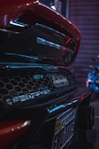 Close-up of the rear section of a Lamborghini car showcasing its distinctive logo and mesh design. Neon lights create reflections on the car's glossy surface, adding a sense of luxury and modernity. The scene is set in an urban environment with motion blur suggesting movement or nighttime setting.