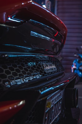 Close-up of the rear section of a Lamborghini car showcasing its distinctive logo and mesh design. Neon lights create reflections on the car's glossy surface, adding a sense of luxury and modernity. The scene is set in an urban environment with motion blur suggesting movement or nighttime setting.